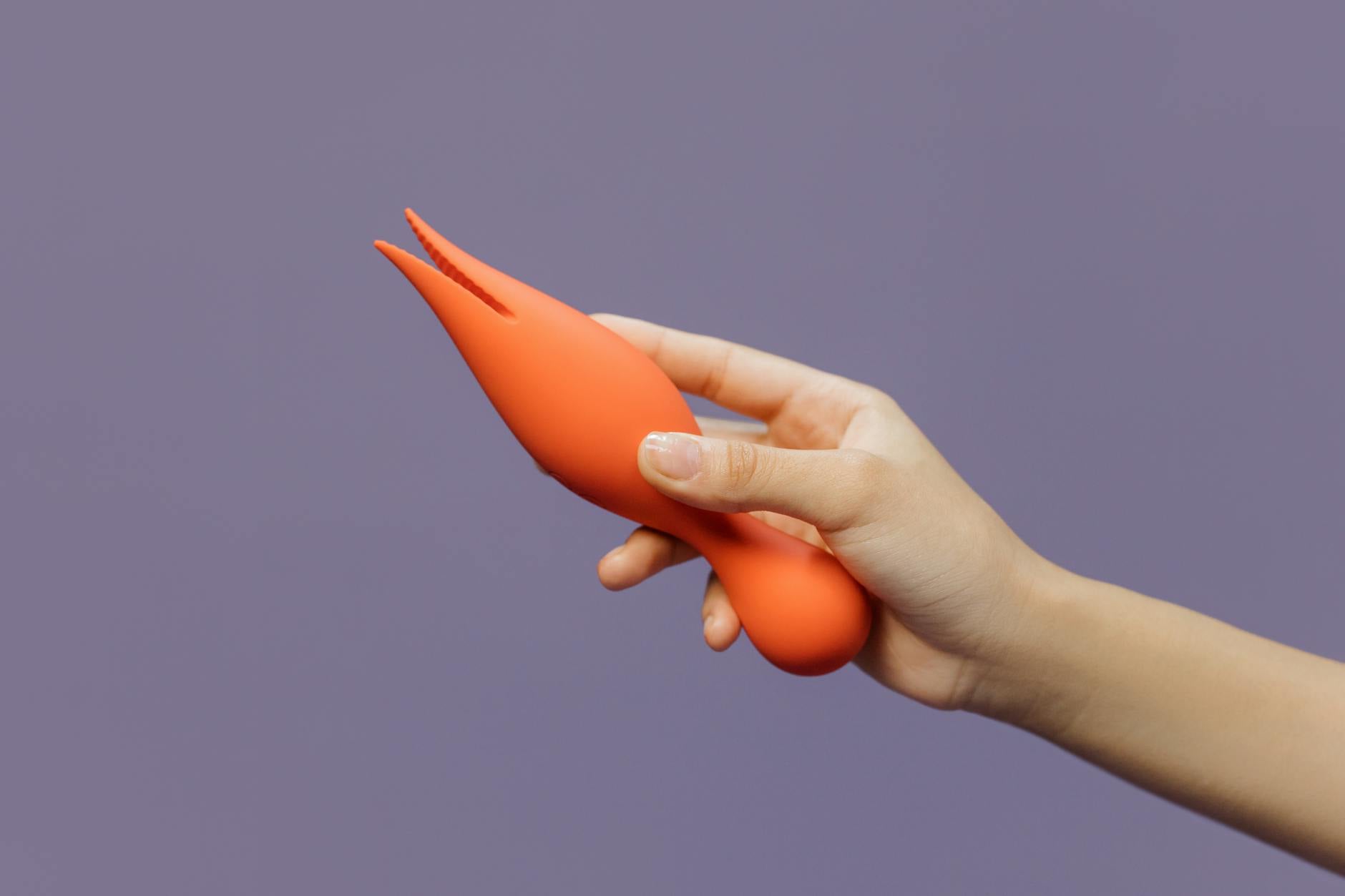 A hand holding an orange vibrator against a minimalistic purple backdrop