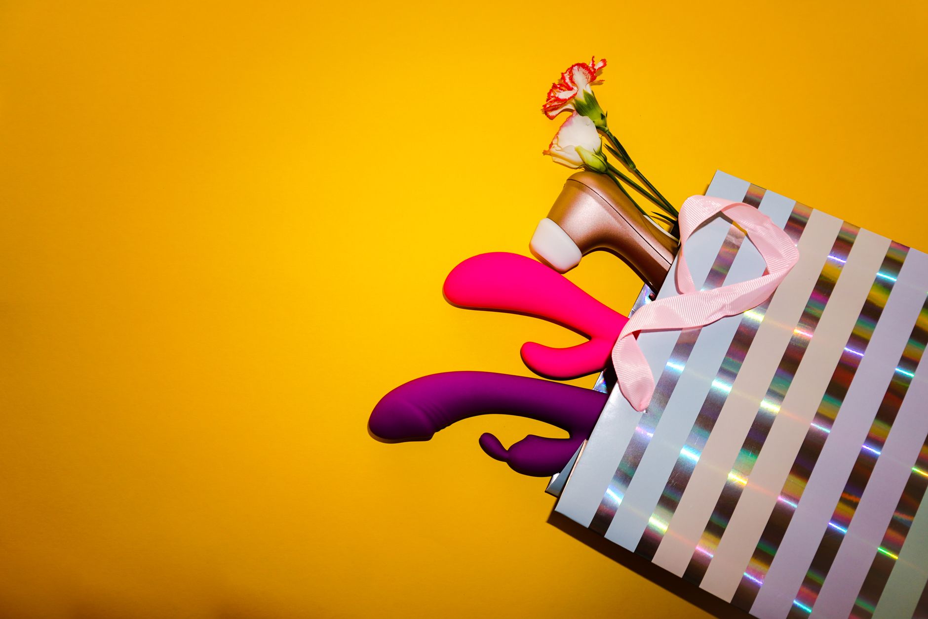 Colorful vibrators with flowers in a holographic gift bag on a yellow background